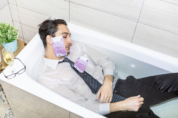 Stressful businessman lying in the bath full of water with banknotes and trying to control his...