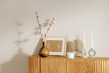 A wooden chest of drawers in a modern interior stands against a white wall. Home decor paraphernalia candles, a photo frame and a vase stand on a chest of drawers in the living room.
