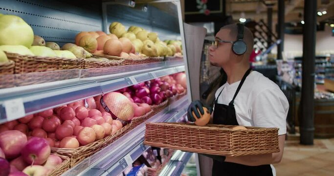 Man worker in black apron and gloves stocking the fruits in supermarket while listen to the music