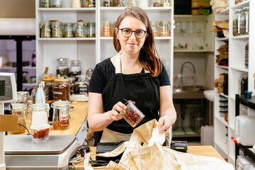 Young friendly confident woman in eyeglasses wearing black apron puts groceries, goji berry...