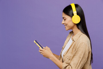 Young happy fun latin woman wear light shirt casual clothes yellow headphones listen to music hold use mobile cell phone isolated on plaint pastel purple background studio portrait. Lifestyle concept.