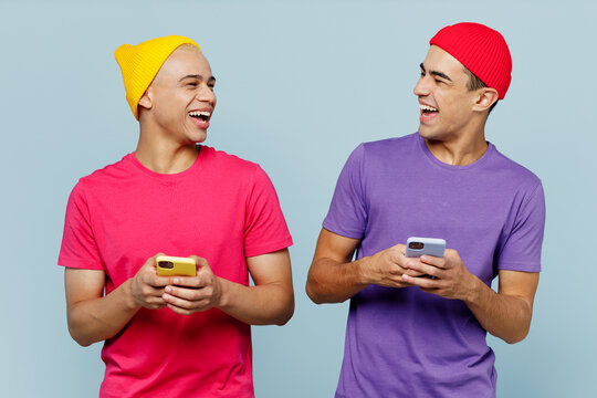 Young cheerful fun couple two friends men wear casual clothes together hold in hand use mobile cell phone look to each other laugh isolated on pastel plain light blue cyan background studio portrait.