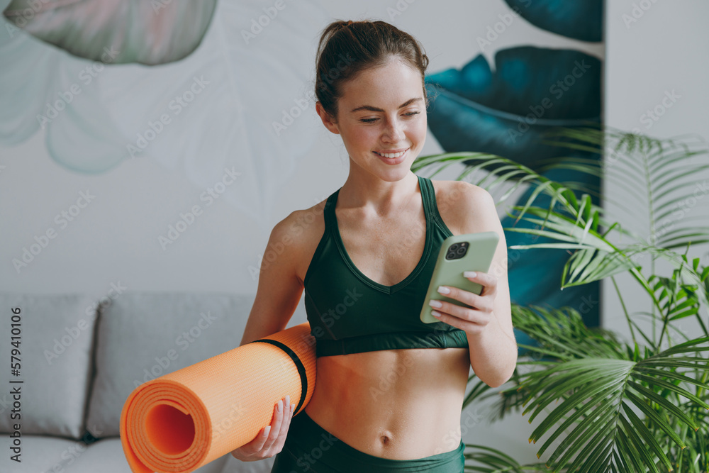 Wall mural Young sporty athletic fitness trainer instructor woman wearing green tracksuit hold in hand yoga mat use mobile cell phone training do exercises at home gym indoor. Workout sport motivation concept.