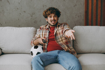 Young sad man fan wearing casual clothes cheer up support football team spread hand switch channels sit on grey sofa with soccer ball rest watch tv indoors room gray color wall Sport leisure concept.