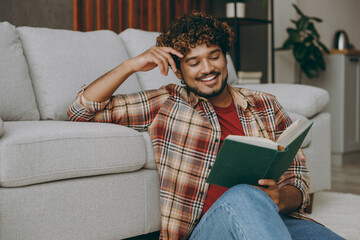 Young smiling happy cheerful Indian man wears casual clothes reading book novel sits near grey sofa...