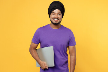 Fun devotee Sikh Indian IT man ties his traditional turban dastar wear purple t-shirt hold use work on laptop pc computer isolated on plain yellow background studio portrait. People lifestyle concept.