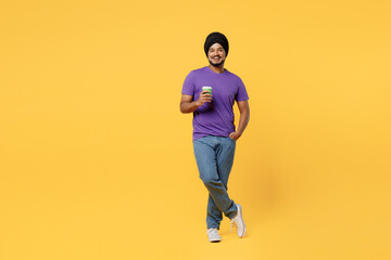 Fototapeta na wymiar Full body devotee Sikh Indian man ties his traditional turban dastar wear purple t-shirt hold takeaway delivery craft paper brown cup coffee to go isolated on plain yellow background studio portrait.