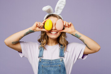 Young smiling cheerful fun woman wears casual clothes bunny rabbit ears cover eyes with colorful eggs isolated on plain pastel light purple background studio portrait. Lifestyle Happy Easter concept.