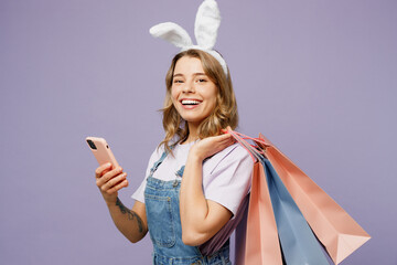 Naklejka na ściany i meble Young woman wear casual clothes bunny rabbit ears hold paper package bag after shopping use mobile cell phone isolated on plain light pastel purple background studio Happy Easter sale buy day concept