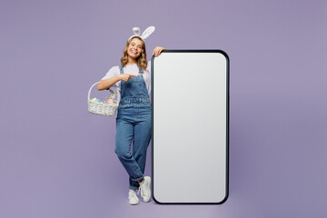 Full body young woman wear casual clothes bunny rabbit ears point index finger indicate on big huge blank screen mobile cell phone with area isolated on plain purple background. Happy Easter concept. - obrazy, fototapety, plakaty