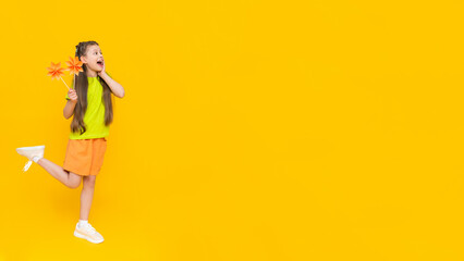 A little girl holds origami flowers on a stick and stands tall. Children's creativity. A child in shorts and a T-shirt dreams of a sunny summer on a yellow isolated background. Banner. Copy space.