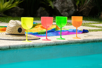 Na borda da piscina copos de drinks coloridos com um chapéu de palha e canga sobre a grama e ao...