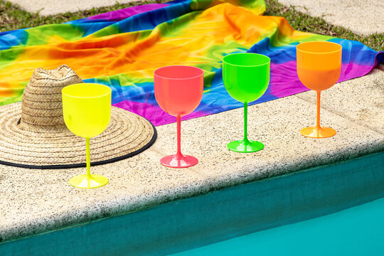 ta&ccedil;as de drink coloridas de amarela, vermelha, verde e laranja na beira da piscina, com um chap&eacute;u de palha e uma canga colorida sobre a grama.