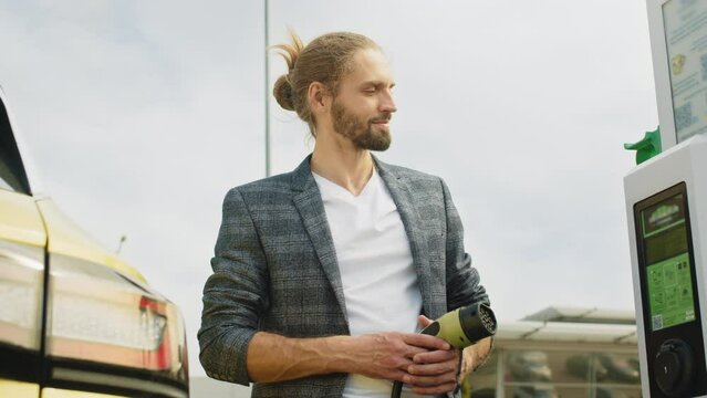 Eco and clean energy concept. An eco urban businessman is leaning on his electic vehice dressed in a plaid suit, man use the white power cable and plug. Port plugging in EV modern car