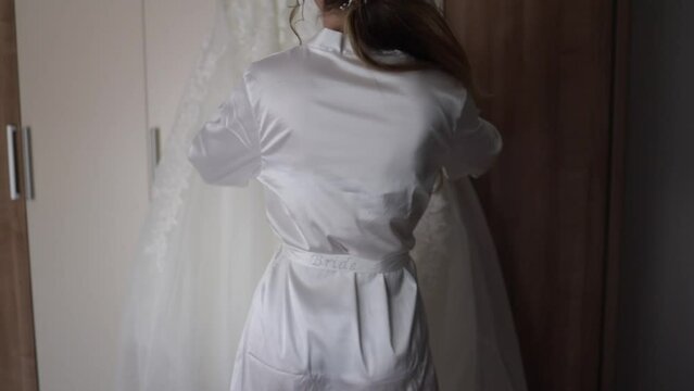 Rear shot of  a bride in a white satin robe touching her wedding dress in a room