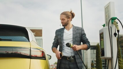 A young European man dressed in casual clothes connects a power cable to an electric car. The video was filmed in daylight. Smart dressed Business man charging electric car at charging station.