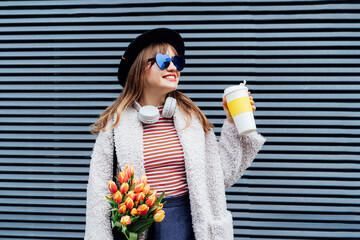 Happy young fashion woman in heart shaped sunglasses, headphones holding coffee cup and netbag with fresh tulip flowers on the gray striped wall background. Urban city street fashion. Spring mood.