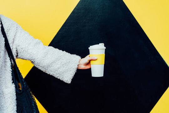 Woman's hand holding reusable white coffee cup on the geometry bright yellow and black wall background. Plastic free, eco-friendly, zero-waste lifestyle concept. Selective focus, copy space