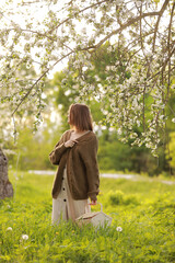 Beautiful young woman wearing dress and khaki knitted cardigan, holding wicker basket in spring in a blooming apple garden in sunny weather. The concept of youth, love, fashion and lifestyle.