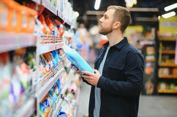 Young man in the supermarket in the household chemicals department. Large selection of products. A brunette in a glasses and a beard in a beige coat.