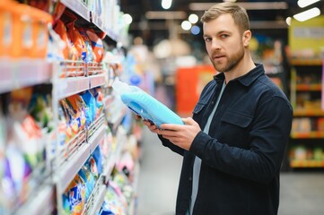 Young man in the supermarket in the household chemicals department. Large selection of products. A brunette in a glasses and a beard in a beige coat.