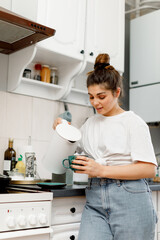 A brunette girl with hair styled in a bun is making tea in the kitchen in the morning. A girl in a white T-shirt and jeans is making tea in a white kitchen.