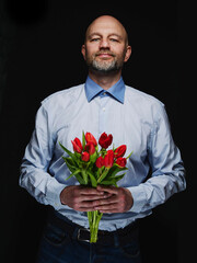 Bald man in blue shirt holding bouquet of red tulips. Selective focus. Male in his 40s with grey beard giving a present to his woman. Dark background.