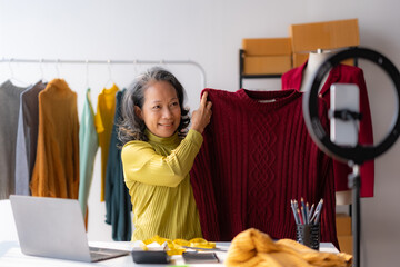 Asian senior female online seller reviewing shirt video live streaming to social media at home.