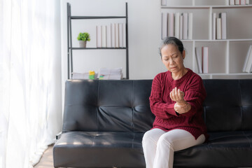 Asian old woman with body aches, leg pain, bone pain, stiff muscles, arm pain, hand pain, sitting on the sofa in the living room stretching to relax.