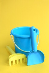 A blue children's bucket with a shovel and a rake for playing is on a yellow background	
