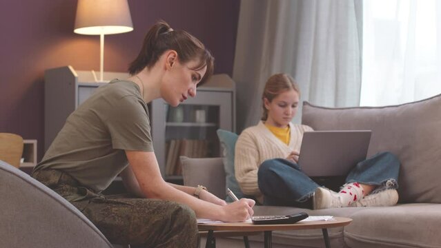 Young Caucasian military woman counting army budget working from home while her preteen daughter using laptop on couch in cozy living room