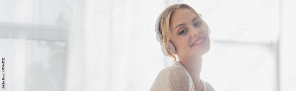 Wall mural overjoyed young woman listening music in wireless headphones while smiling at home, banner.