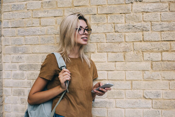 Fototapeta premium Smiling woman with handbag and smartphone standing near brick wall