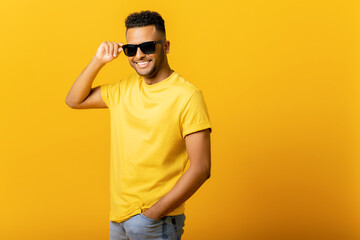 Overjoyed Indian man wearing sunglasses looking at the camera, smiling positively, being in high spirit. Carefree hispanic guy in yellow t-shirt isolated, having summer mood - Powered by Adobe
