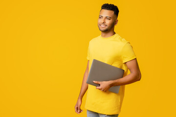 Cheerful handsome indian male student holding laptop standing isolated on plain yellow background, man in casual yellow wear advertising, recommending new computer app