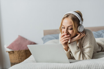 cheerful woman in wireless headphones watching comedy movie on laptop in bedroom.