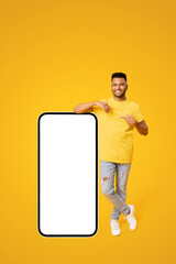 Look here. Carefree happy Indian 20s guy standing and leaning on giant huge smartphone with empty...