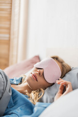 blonde woman in pink sleeping mask and blue pajama resting in bed.