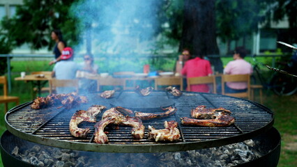 Person preparing meat on grill outside with friends and family in background. Closeup hand holding clamp cooking food at BBQ party