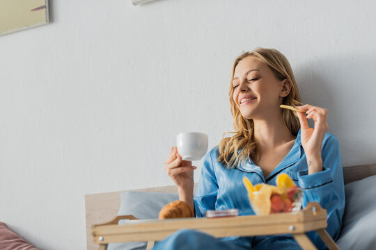 Joyful Woman In Pajama Holding Cup Of Coffee And Dried Mango Near Tray While Having Breakfast In Bed.