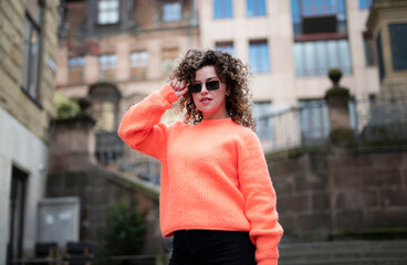 Fashionable young woman with curly hair in orange sweater and dark sunglasses walking in the sunny city