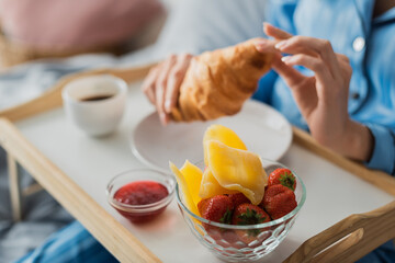 cropped view of woman holding fresh croissant near tray with jam and dried mango while having breakfast in bed.