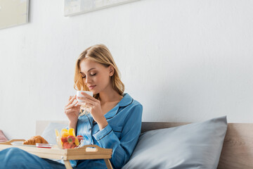 pretty woman holding cup while enjoying coffee flavor near breakfast tray in bed.