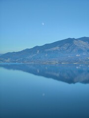 Vertical aerial view of a beautiful lake reflecting the mountain with the Moon against a blue sky