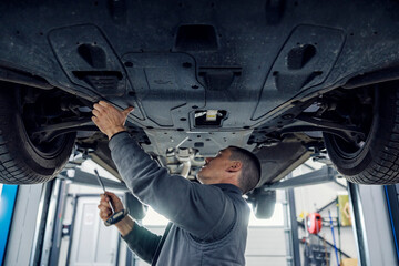 An auto mechanic is repairing car while standing under it at mechanic's shop. - obrazy, fototapety, plakaty
