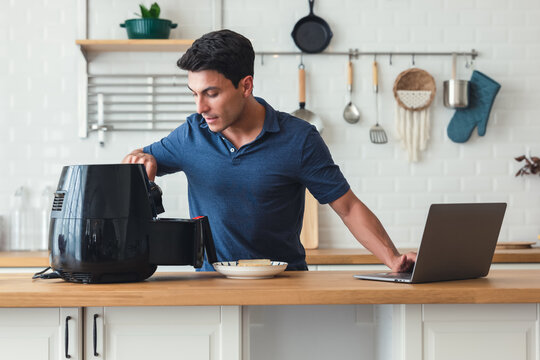 Handsome Man Standing At Kitchen Table And Cooking Toast Bread Healthy With Recipe By Air Fryer Machine, Using Laptop Computer Learning Online Cooking Class In Modern Kitchen At Home.