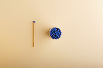 Top view of one long wooden match with a blue head next to an open round box with a whole set of such matches, on a light yellow background - obrazy, fototapety, plakaty