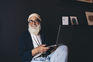 Cheerful hipster senior man sitting on chair with laptop