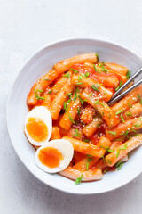 Tteokbokki with eggs, fresh green leek and  roasted sesame seeds in blue  bowl on the white  table top. Tteok-bokki is a   Korean cuisine dish with rice gnocchi sticks and spacy sauce.  Asian food
