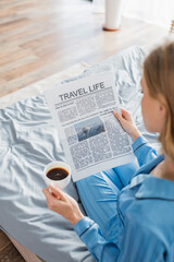 overhead view of blonde woman reading travel life newspaper and holding cup of coffee in bedroom.